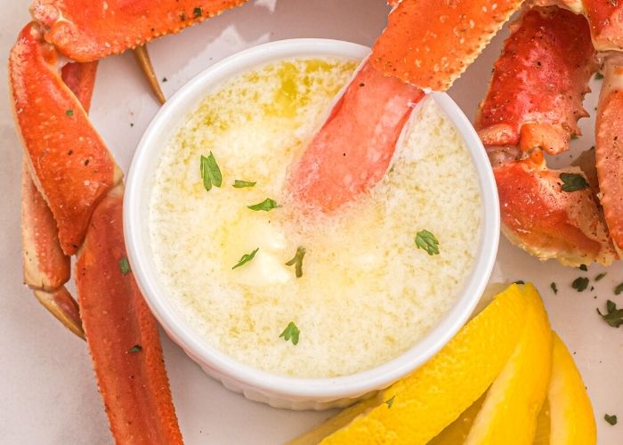 Crab leg meat, being dipped into melted butter, served on a white plate with parsley flakes garnished on top. 