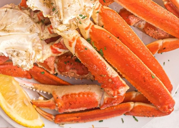 Juicy crab legs, orange in color, with lemon slices served on a white plate with parsley flakes. 