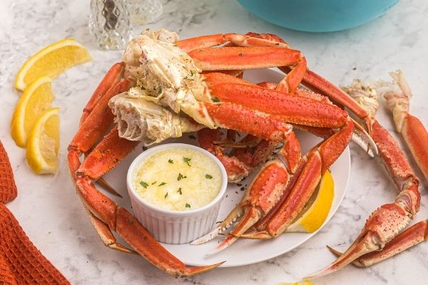 Moist and orange crab legs, on a white plate, served with lemon slices and melted butter on a marble table. 
