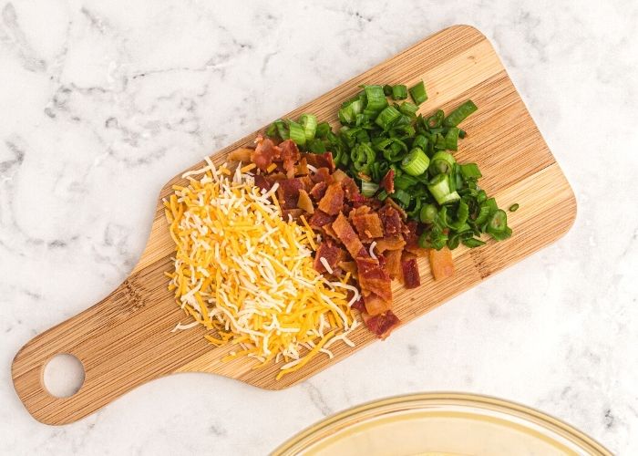 Shredded cheese, chopped green onions, and crumbled cooked bacon and a cutting board.