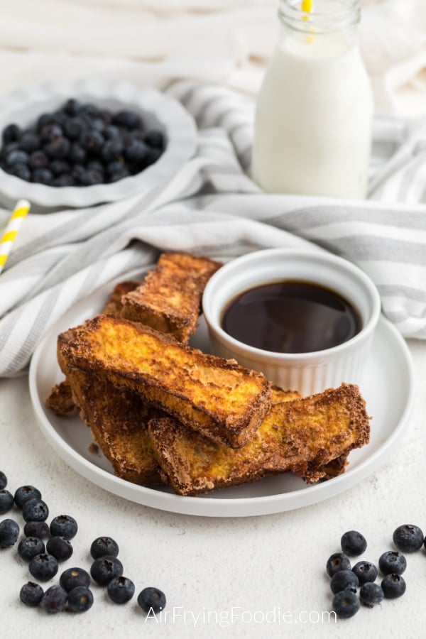 French toast sticks that were made in the Air Fryer on a white plate with a small bowl of syrup and surrounded by blueberries. 