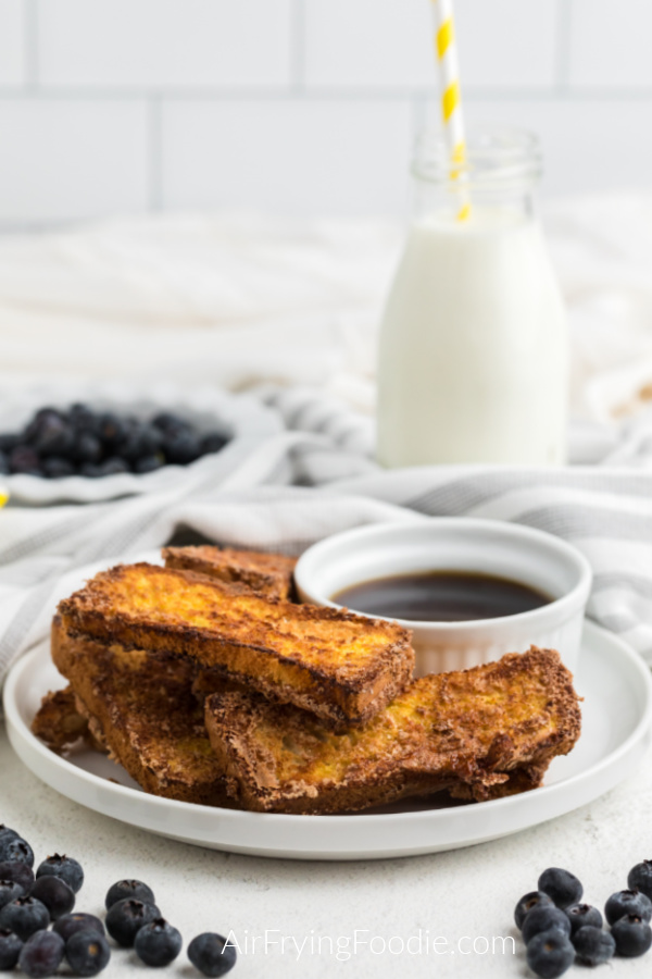 French toast sticks that were made in the Air Fryer on a white plate with a small bowl of syrup and surrounded by blueberries. 