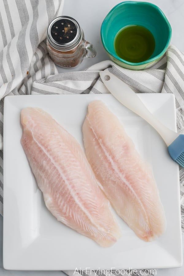 uncooked swai fillets in a white plate with a brush, and blackened seasoning and olive oil above. 