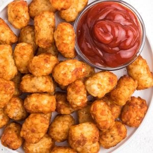 Tater Tots on a white plate that were made in the Air Fryer. A small bowl of ketchup on the side.