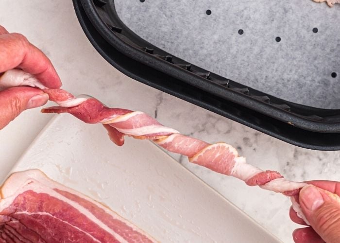 Raw bacon being twisted lengthwise, and then place into the air fryer basket, lined with parchment paper with holes. 
