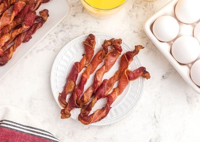 Five strips of crispy twisted bacon on a white plate with a tray of eggs next to it.
