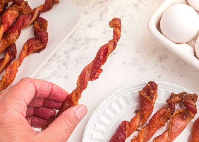 A hand holding a piece of twisted bacon over a plate of bacon on a white marble table. 