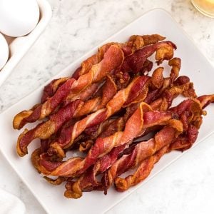 Crispy twisted bacon, stacked and served on a white square plate, next to orange juice and eggs.