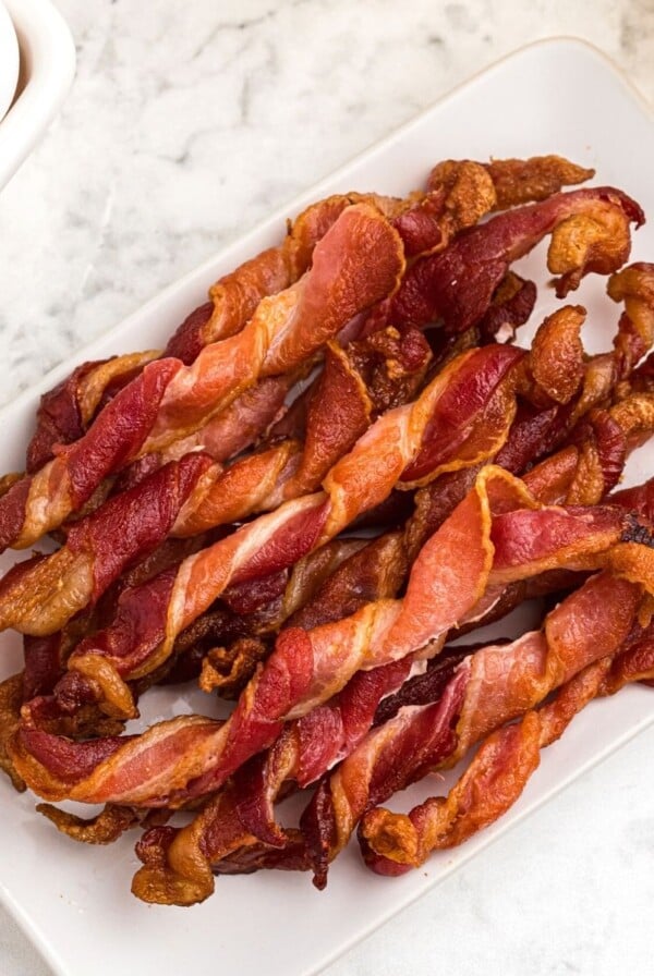 Crispy twisted bacon, stacked and served on a white square plate, next to orange juice and eggs.