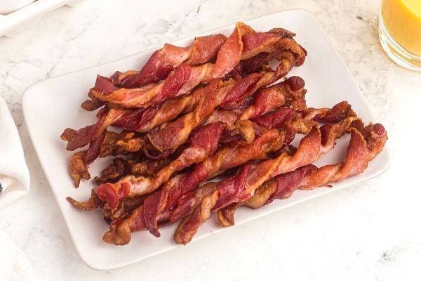 Crispy bacon twisted into curls and stacked on a white rectangle plate. Orange juice in a glass on the side. 
