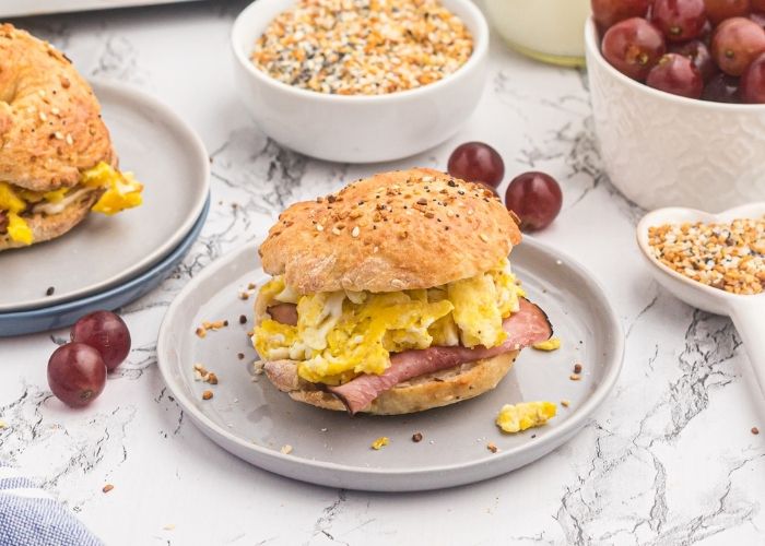 Air fryer bagel served with ham and eggs on a light gray plate on a marble table. Grapes scattered around the table. 