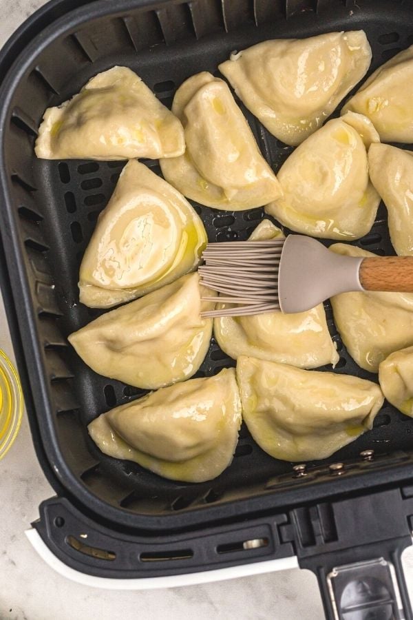 Uncooked pierogies in an air fryer basket, being brushed with olive oil.