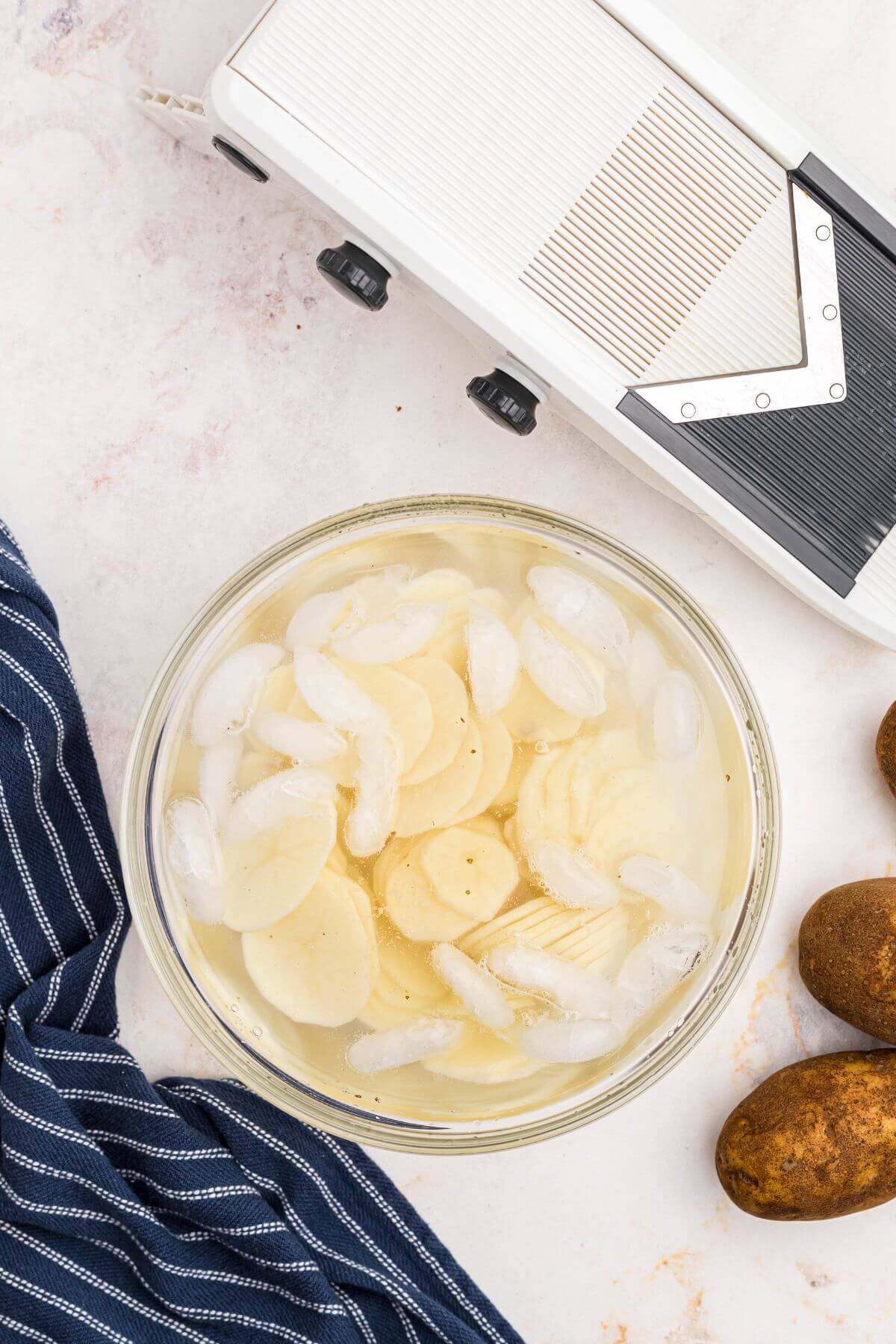 Potatoes cut into slices in front of a mandolin slicer on a white marble table. 