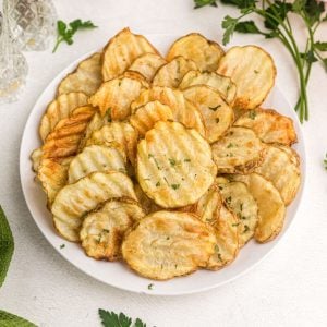 Close up of potato chips on a white plate with fresh parsley garnish on the table.