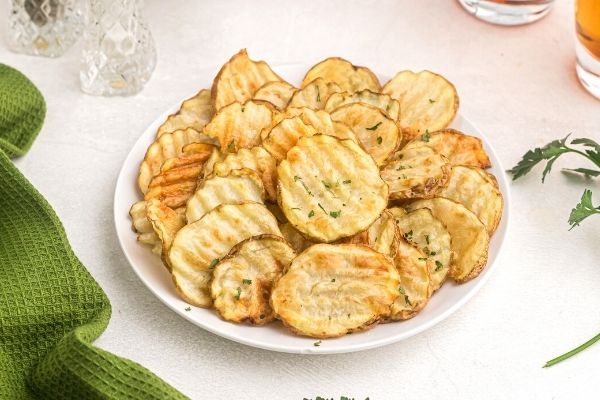 Golden potato chips piled on to a white plate with a green towel and parsley flakes sprinkled on top. 