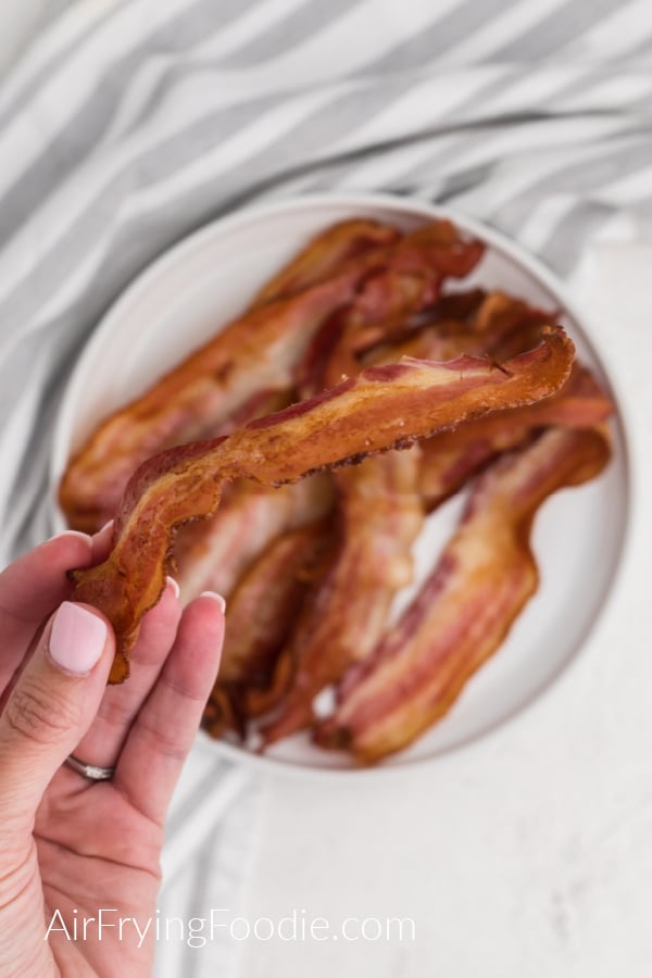 Hand holding crispy bacon over a plate of bacon that was made in the air fryer. 