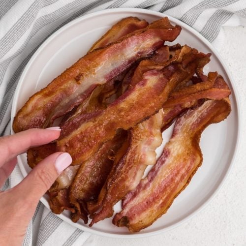 Hand grabbing a piece of air fryer bacon that's on a white plate.