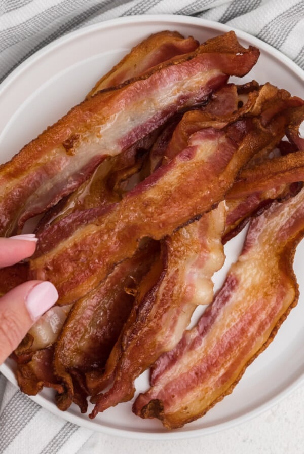 Hand grabbing a piece of air fryer bacon that's on a white plate.