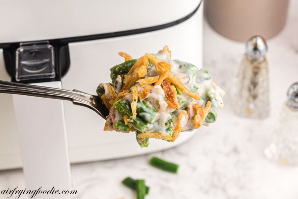 Spoonful of green bean casserole being scooped out of dish in front of air fryer. 