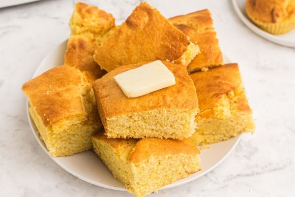 Cornbread cut up into squares and stacked on a white plate, topped with butter. 
