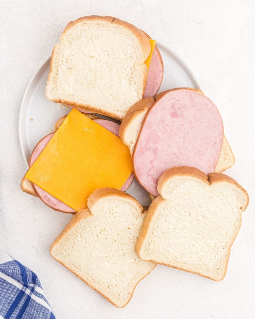 Slices of buttered bread with ham and cheese slices being stacked onto a plate. 