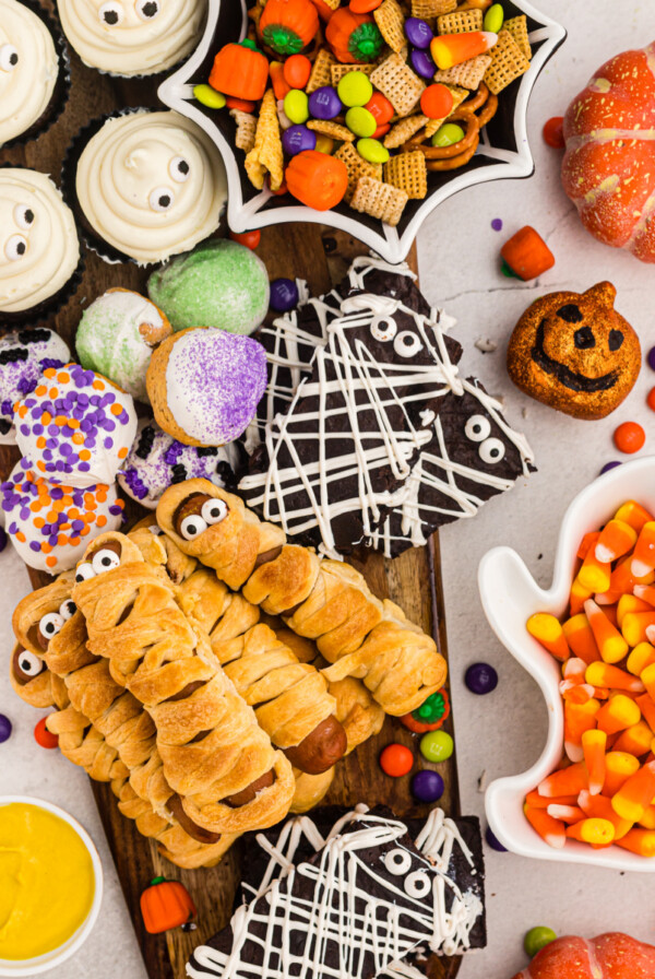 Overhead of board filled with brownies, cupcakes, mummy dogs, donut holes and candy scattered around the table.