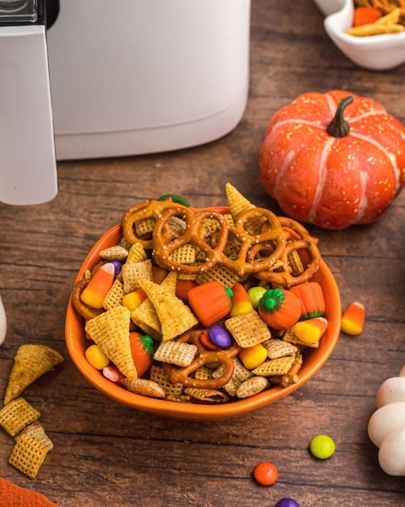 Chex Halloween mixture in an orange pumpkin shaped bowl with candies spread on the table in front of an air fryer. 