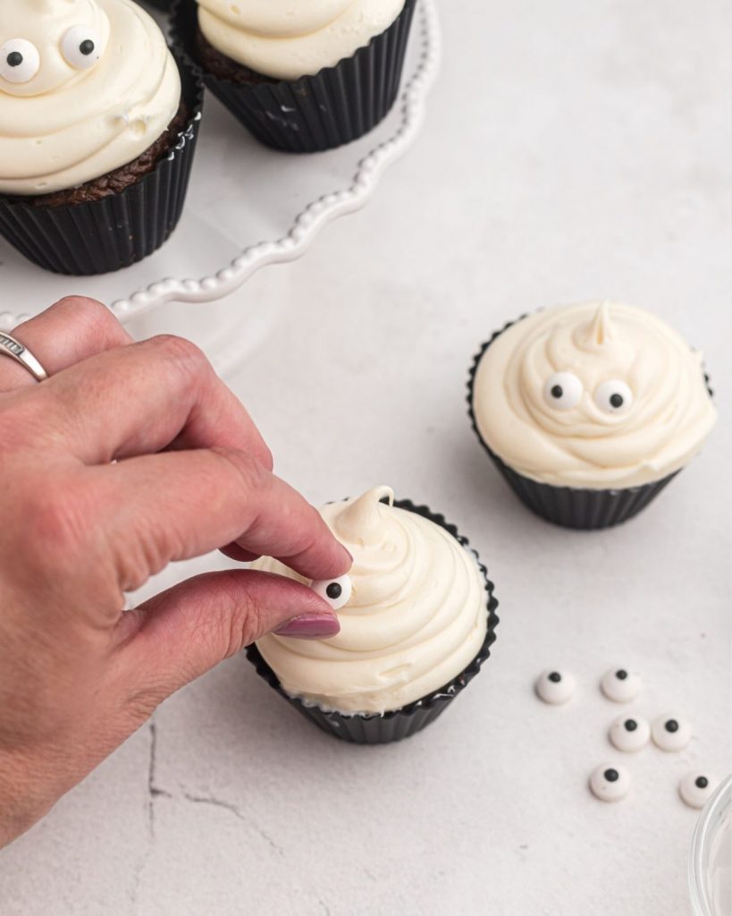 Placing a candy eye on a mound of vanilla frosting on a cupcake with candy eyes scattered on the table. 