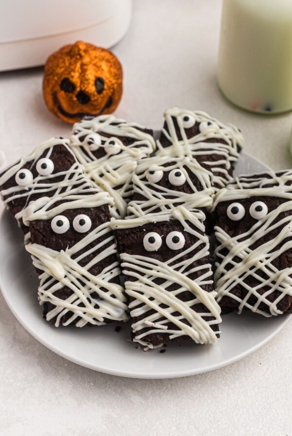 mummy brownies stacked on a white plate with glasses of milk in the back and small Halloween decorations on the table.