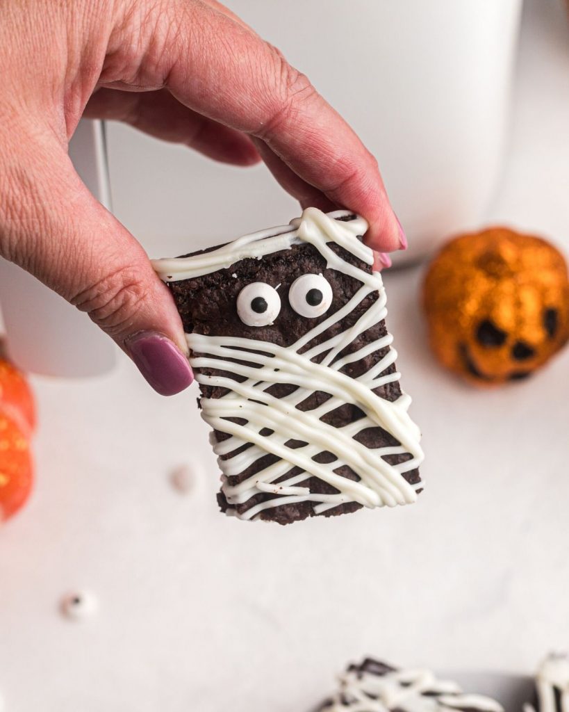 Hand holding brownie mummy with candy eyeballs in front of air fryer. 