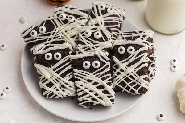 Plate of mummy brownies stacked on a white plate with candy eyes scattered on the table. 