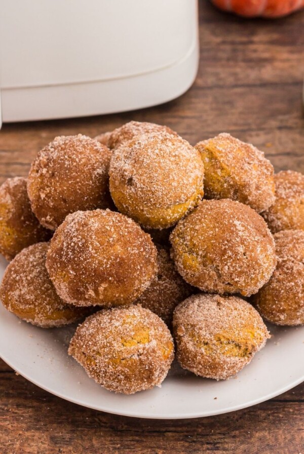 Golden pumpkin donut holes stacked on a white plate in front of glasses of milk and air fryer on a wooden table.