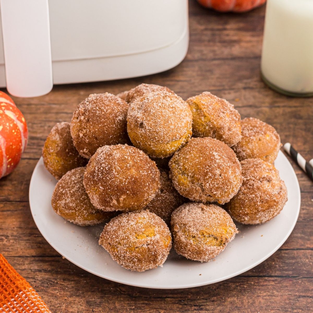 Golden pumpkin donut holes stacked on a white plate in front of glasses of milk and air fryer on a wooden table.