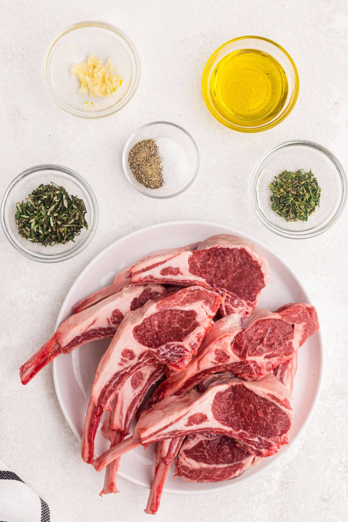 Uncooked lamb chops on a white plate with seasonings on a white table. 