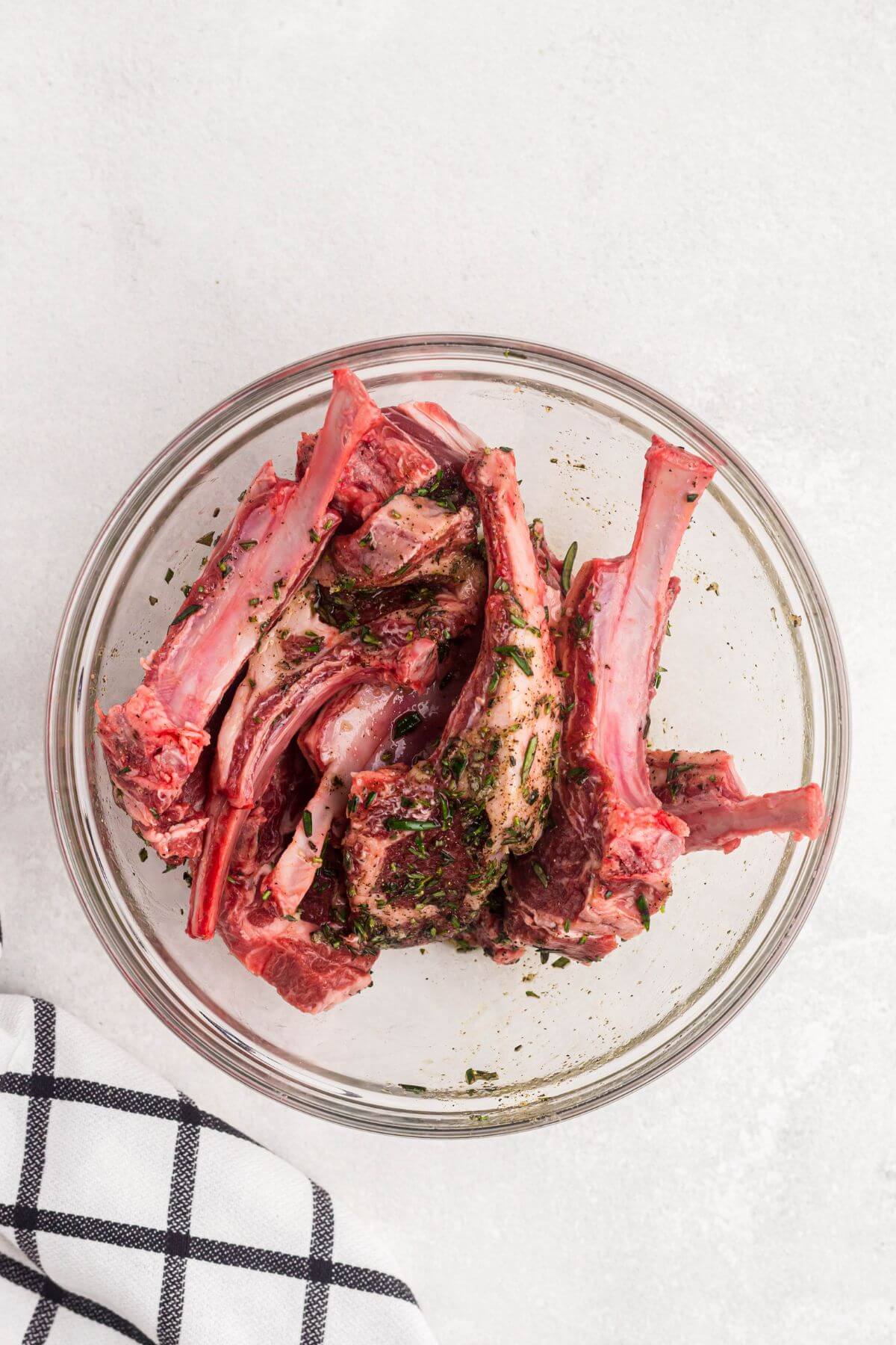 Uncooked lamb chops in a clear glass bowl, tossed with seasonings. 