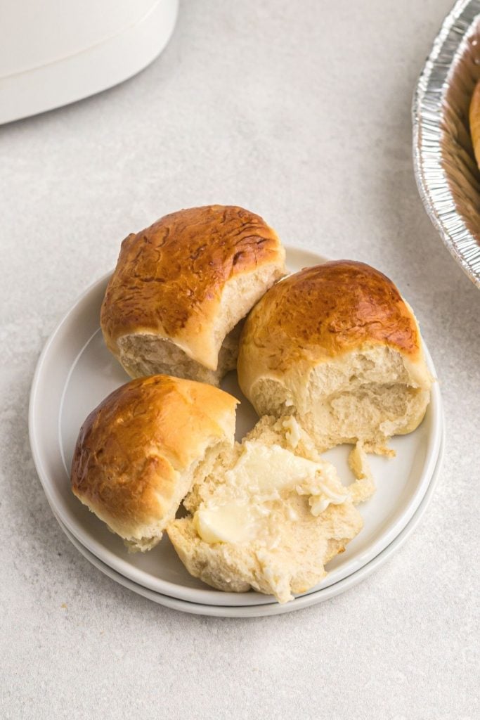 Small rolls on a white plate, cut open and topped with butter. 