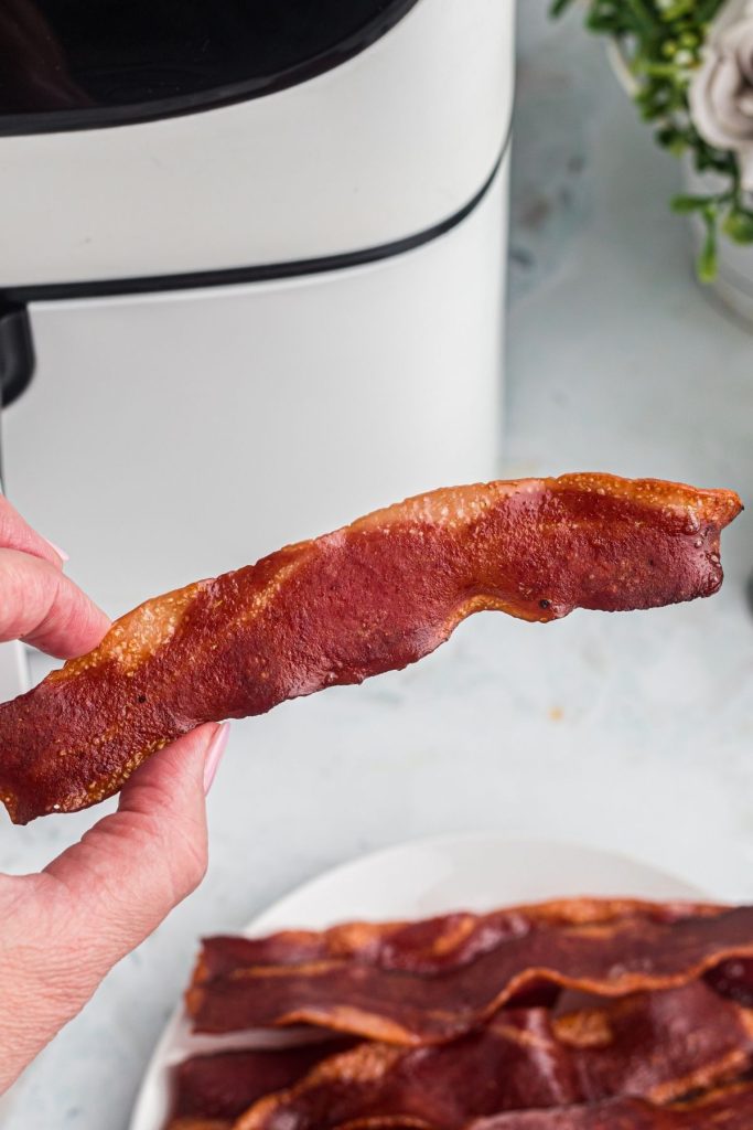 Crispy slice of turkey bacon cooked and being held over the plate. 