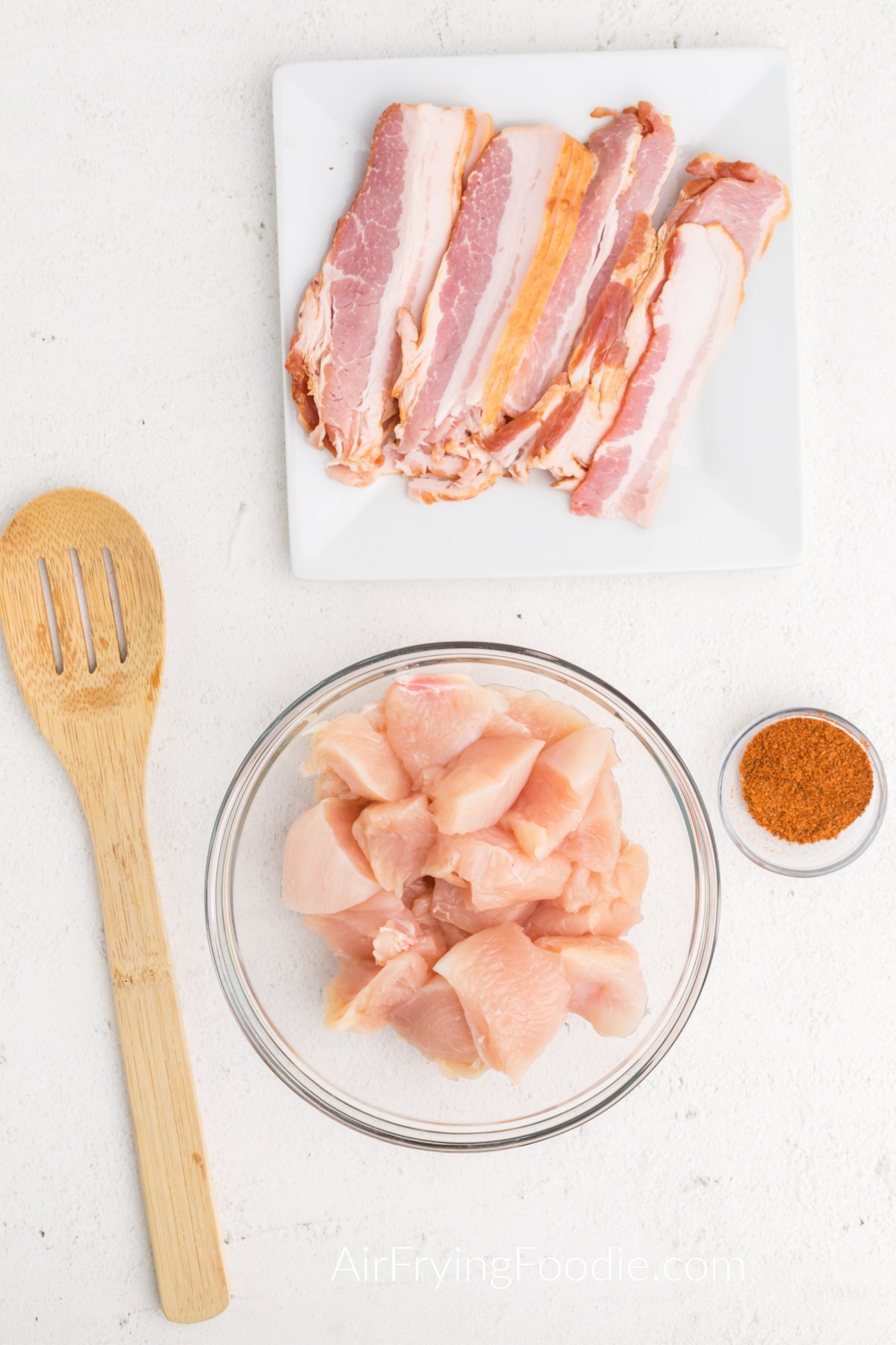 Bacon on a white plate, seasonings in a small container, and chicken pieces in a bowl. 