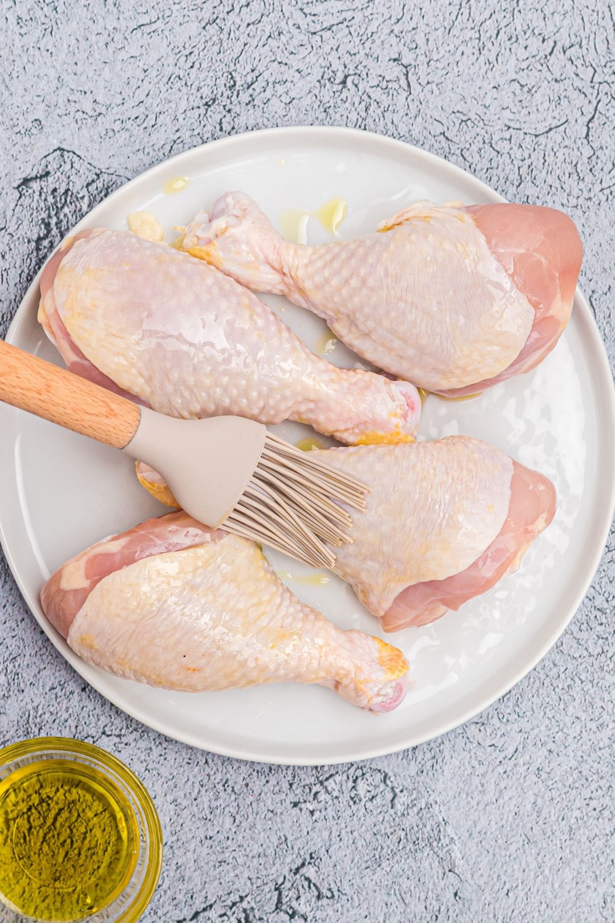 Brushing  uncooked drumsticks with olive oil on a white plate.