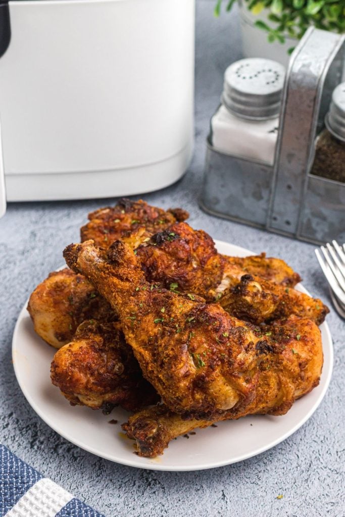 Plate of crispy chicken drumsticks stacked on a white plate on a light blue table in front of an air fryer. 
