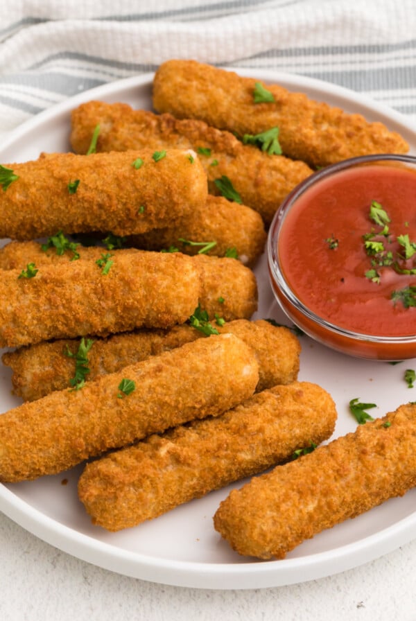 Frozen Air Fryer Mozzarella Sticks on a white plate with dipping sauce and topped with fresh parsley.