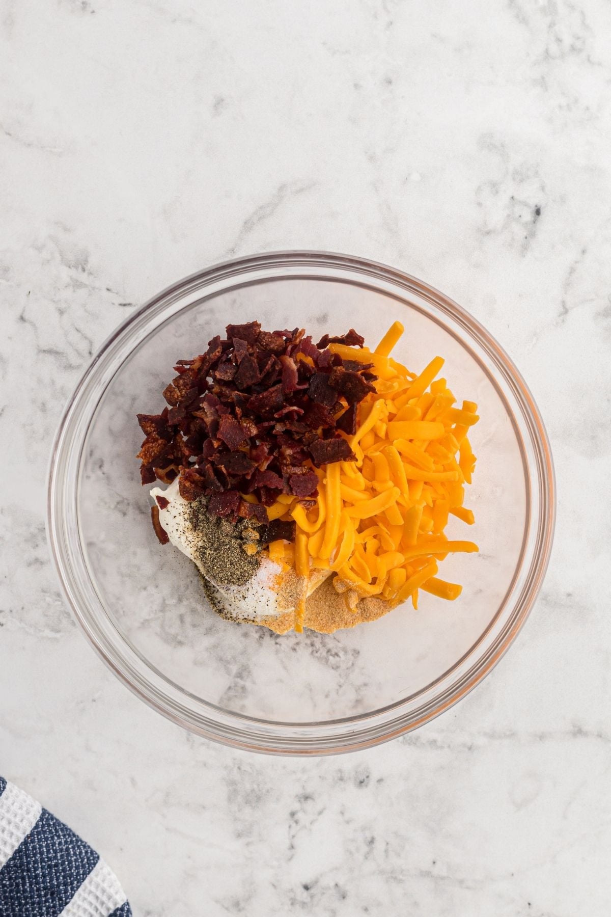Cheese, crumbled bacon, cream cheese, and seasonings in a clear bowl before being mixed