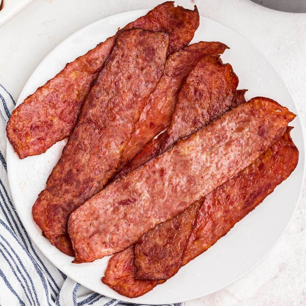 Juicy crispy bacon stacked on a small round plate in front of eggs and air fryer basket.