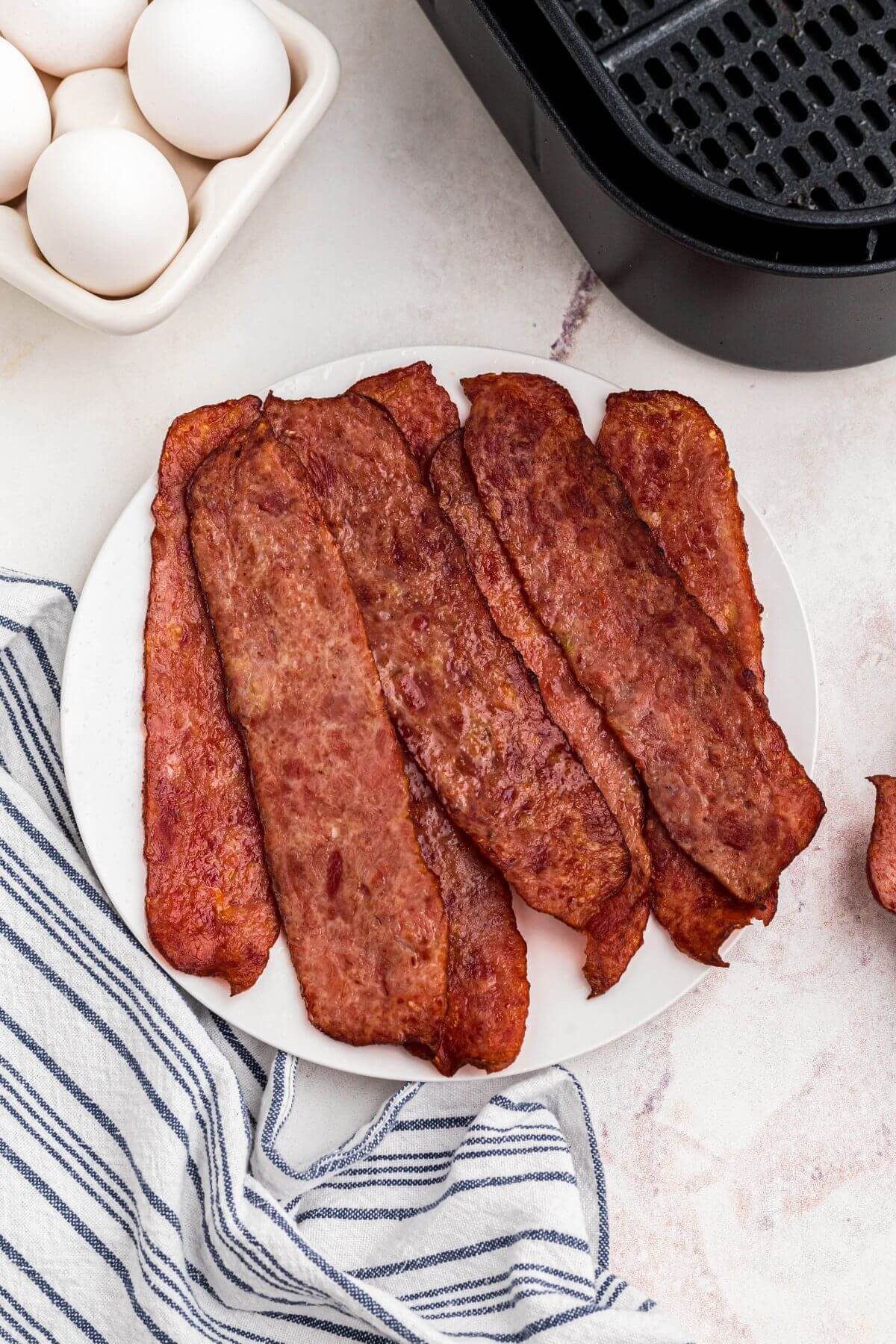 Juicy crispy bacon stacked on a small round plate in front of eggs and air fryer basket. 