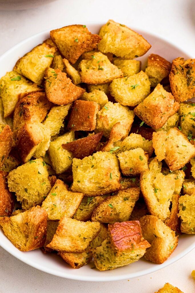 Seasoned golden croutons in a white bowl on a marble table next to a salad.