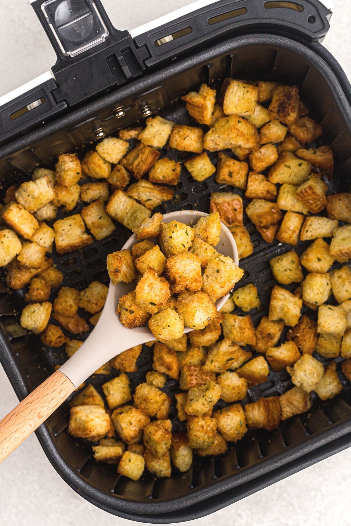 Golden crispy croutons in the air fryer basket being scooped in a serving spoon
