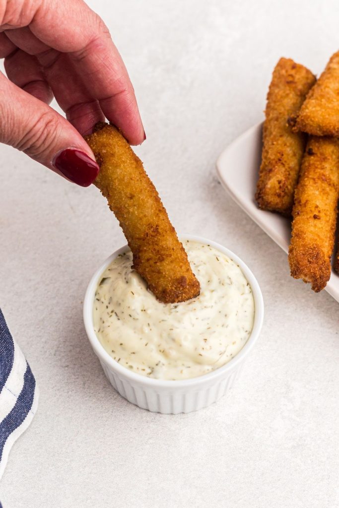 Golden breaded crispy fish stick being dipped into tartar sauce dish.
