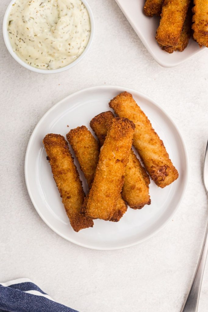 Small white plate with fish sticks served next to large dish filled with fish sticks