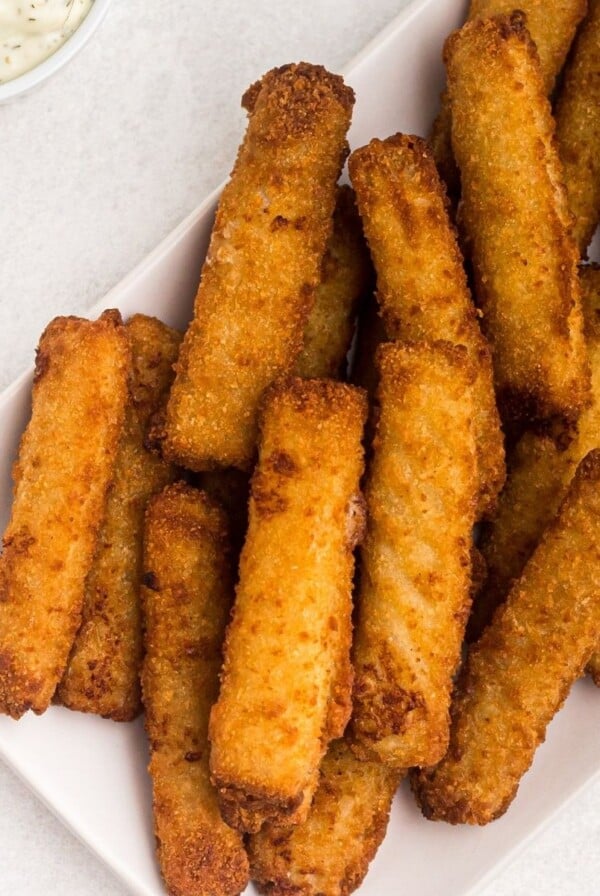 Golden fish sticks stacked on a white plate with a side of tartar sauce