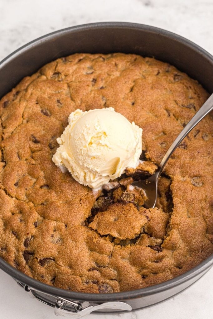 Golden cooked chocolate chip cookie in a pan with a scoop of ice cream and a spoon in the middle
